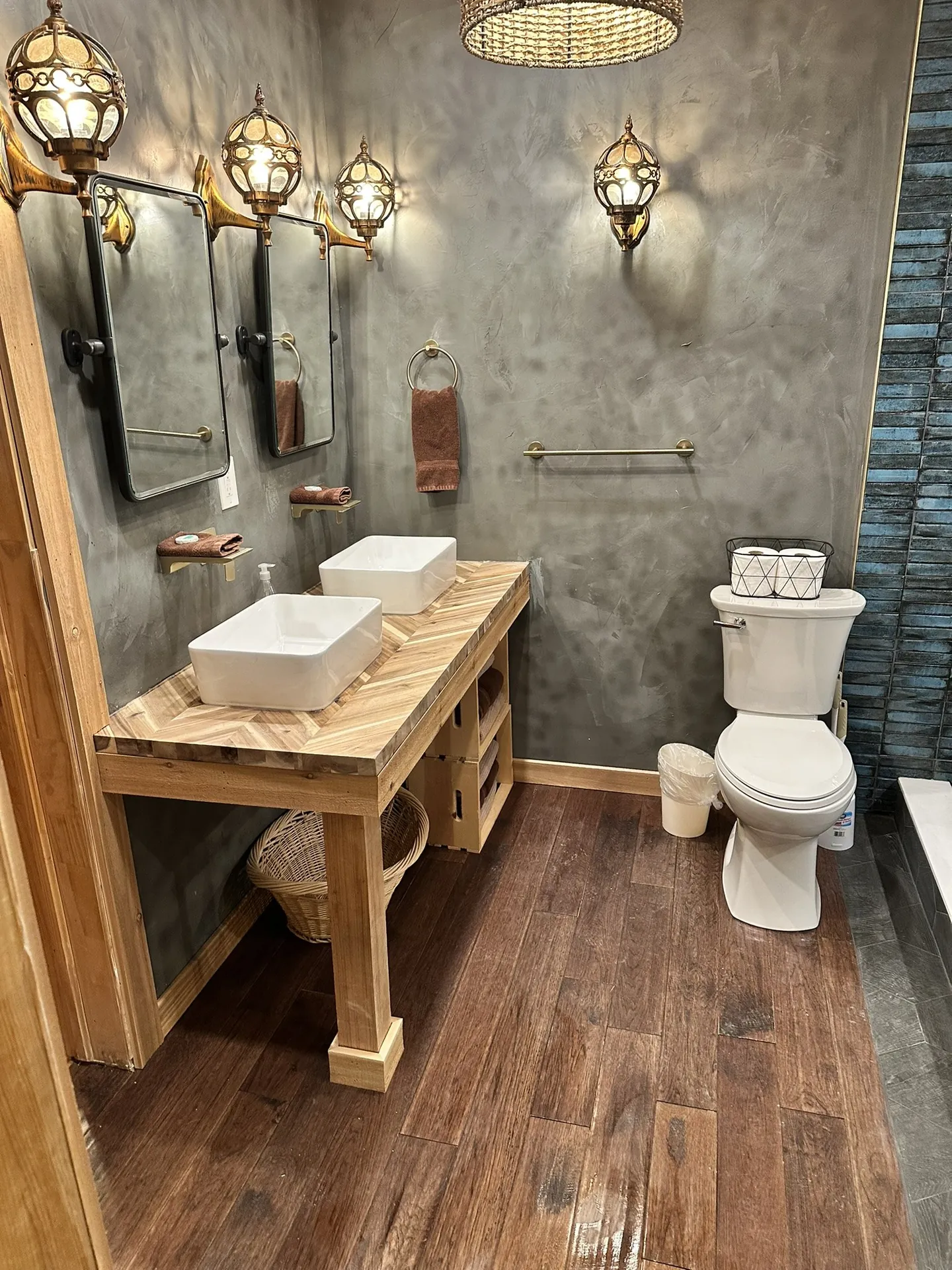 Finished bathroom vanity — double vessel sinks, herringbone wood top, Moroccan brass sconces fully installed, two mirrors, toilet visible