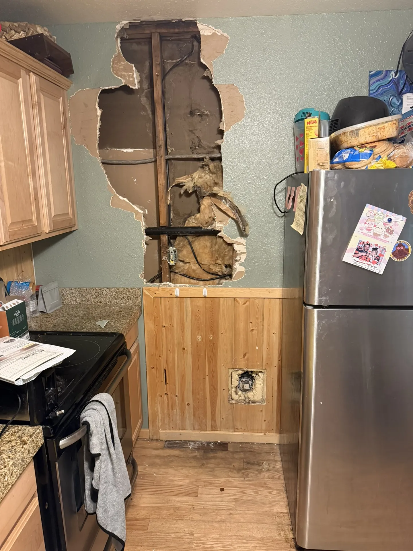 Large hole through the kitchen drywall of a Brian Head cabin exposing studs, insulation, and wiring above knotty pine wainscoting