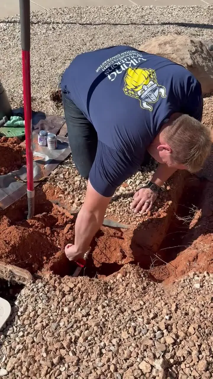 Crew member hand-digging around irrigation lines beside a valve box with PVC primer and cement cans on site