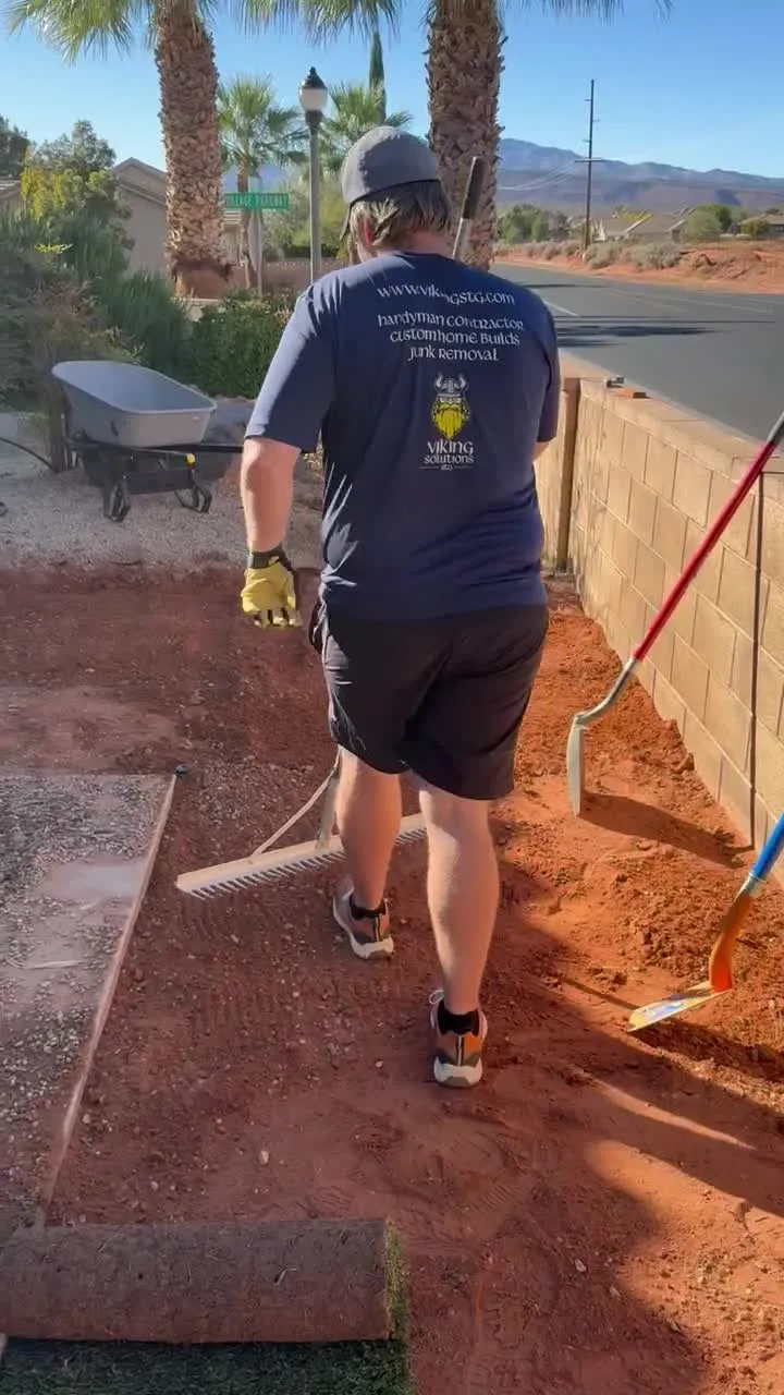 Crew member in a Viking Solutions shirt raking graded soil flat with a sod roll waiting at the edge of the yard